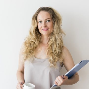 Woman with long blonde hair holding a coffee cup and clipboard, standing against a white background.