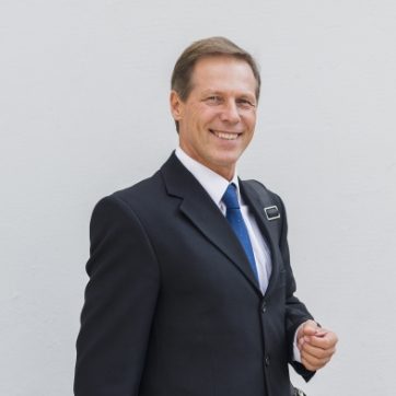 Smiling man in a business suit and blue tie standing against a plain background.