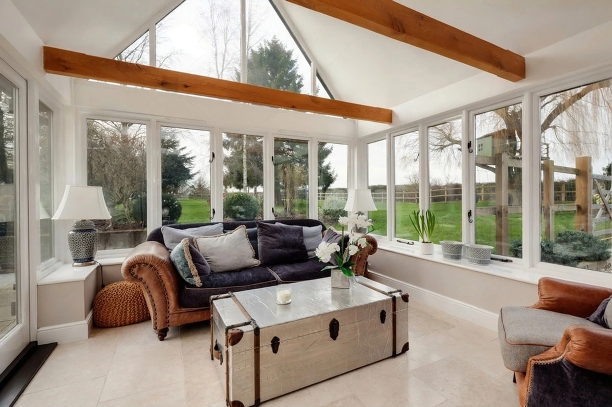 Bright conservatory with exposed beams, large windows, leather sofa, and garden views.