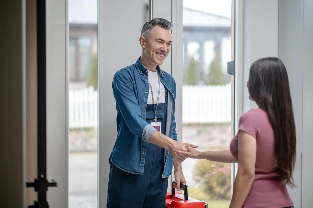Smiling technician greeting a customer at her front door with a handshake and toolkit in hand.