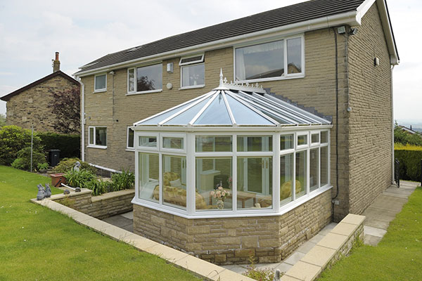 White Victorian-style conservatory with a glass roof attached to a stone house with a sloped garden
